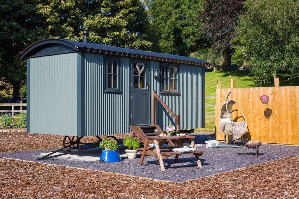 an outdoor view of a Settledown Retreats shepherd hut and the private patio with hanging teardrop chair, patio table and fire pit