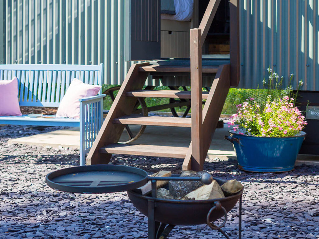 the wooden steps leading up to the luxury shepherd hut - a firepit, enamel flower pot full of wild flowers, and a blue wooden bench on the private patio area out front