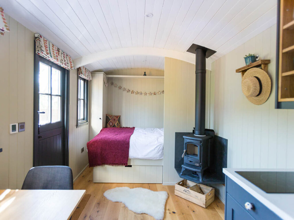 inside the luxury shepherd hut, a dining table and kingsize bed are featured, and a sheepskin rug lies on the floor in front of a woodburning stove. Sun streams through the open door and window