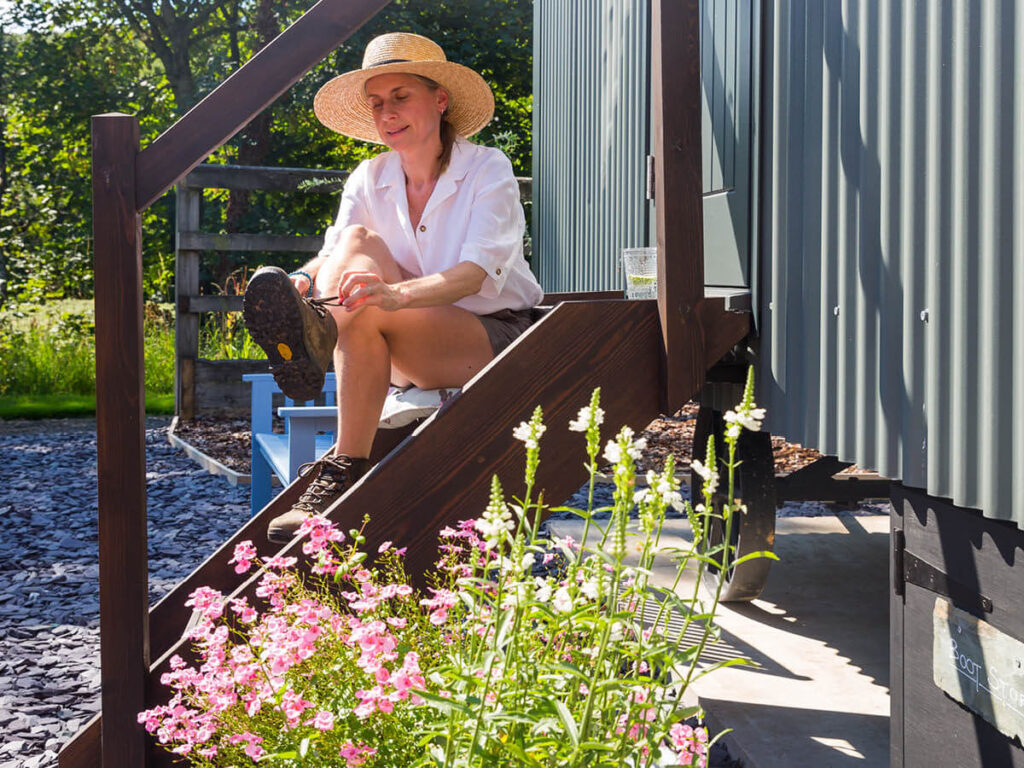 a woman in a straw hat sits on the wooden steps of the shepherd hut tying up her walking boots , beautiful pink wild flowers in the foreground and sun streaming down