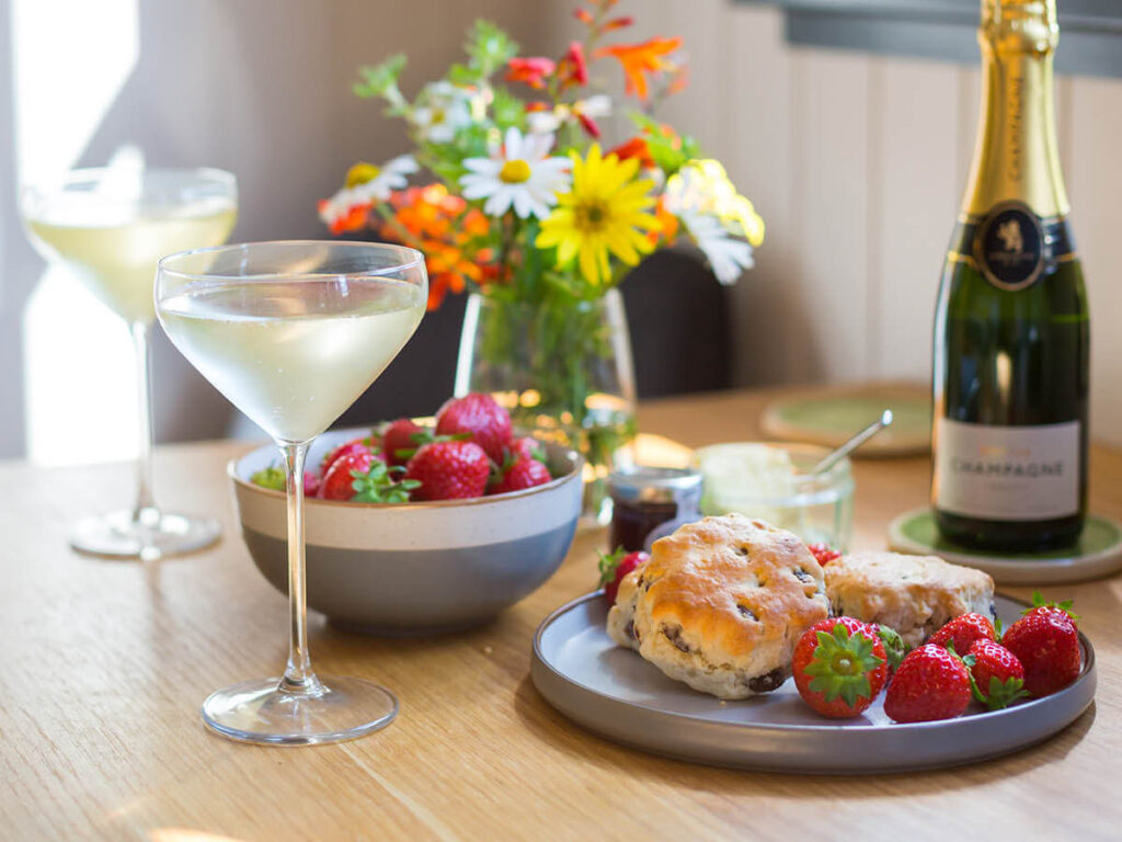 a close up shot of a dining table - on the table is a bottle of Champagne and two full glasses, a bowl of fresh strawberries, a plate with freshly-baked scones and strawberries, and a vase of colourful flowers