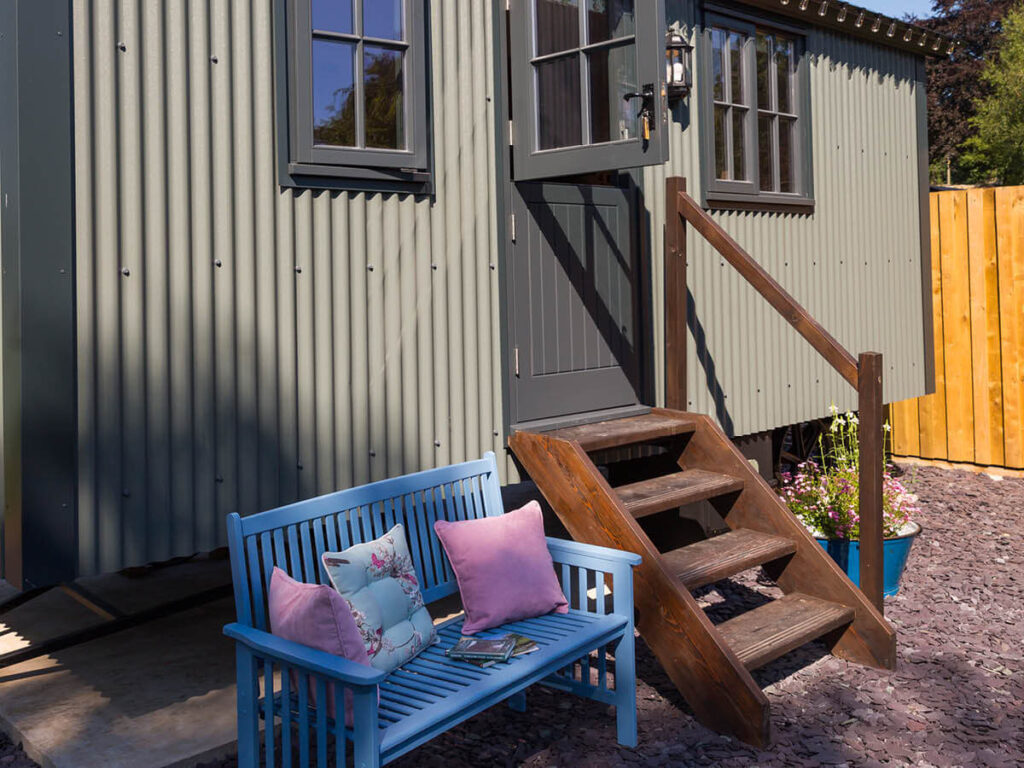 a shepherd hut's with the top half of its stable door swung open, a blue wooden bench with pink cushions and books on sit out front