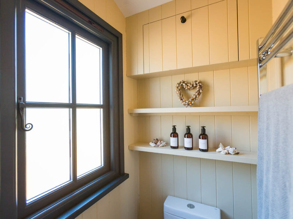 a frosted window in the wood panelled shower room of a Settledown shepherd hut, nice toiletries lined up on a shelf and a decorative heart shaped ornament on the shelf above