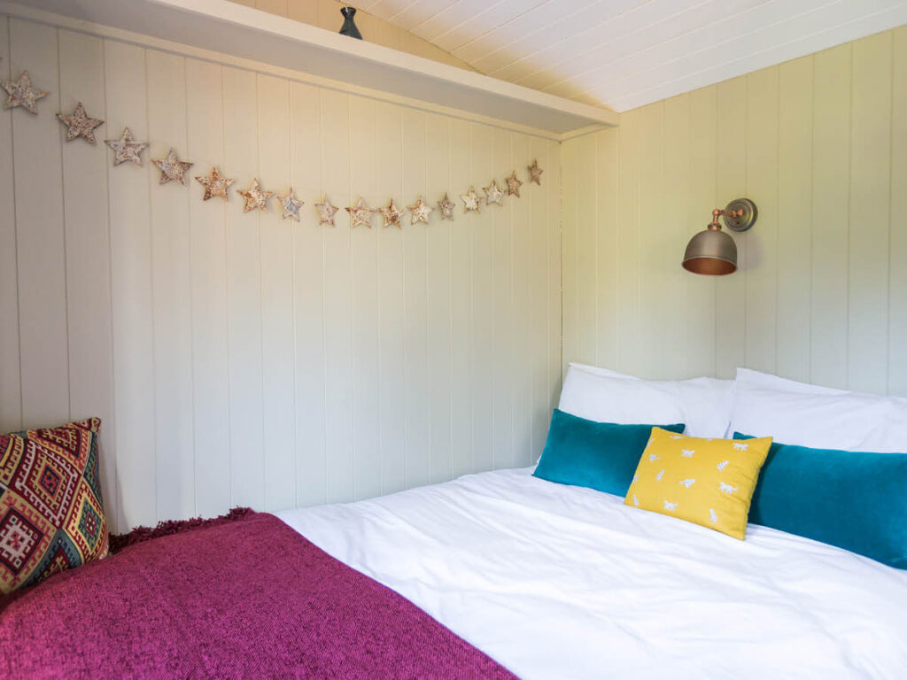 the king size bed inside one of the beautifully furnished shepherd huts at Settledown Retreats, colourful cushions and a throw on top and row of decorative stars hanging on the wall