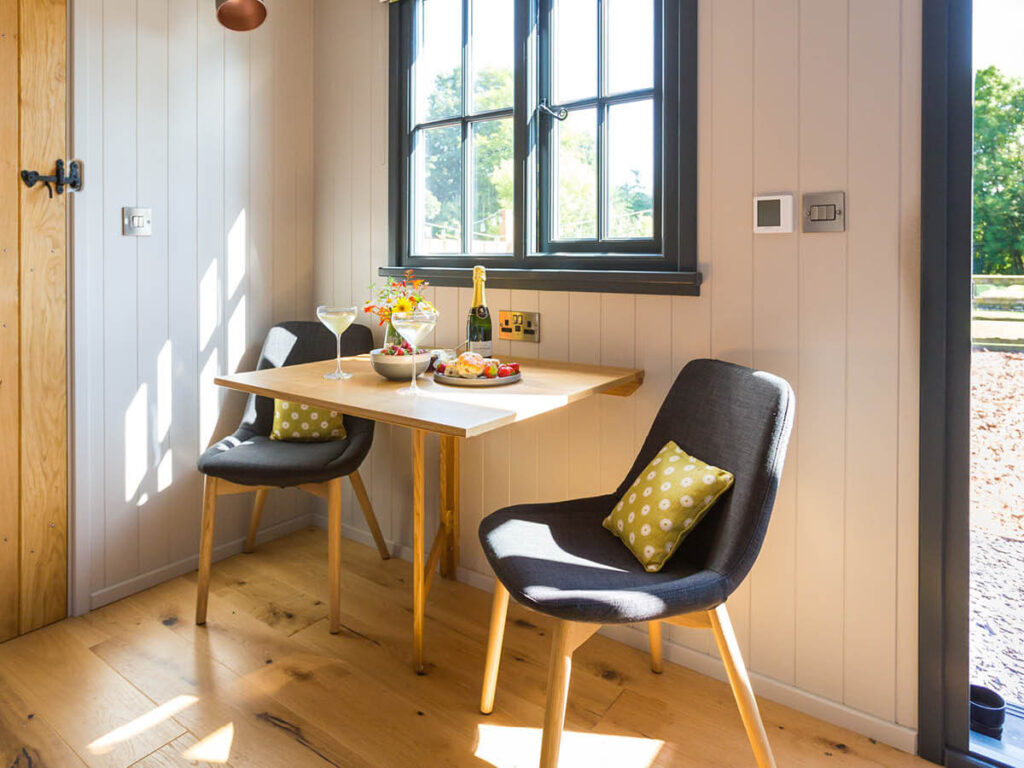 sun streams through the window of a lovely shepherd hut, a bottle of Champagne, food and wild flowers on the table