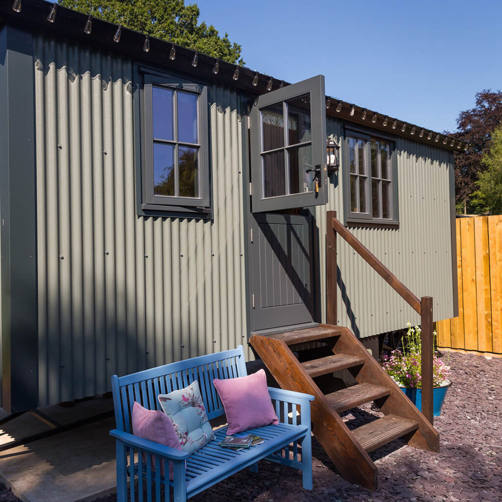 a shepherd hut's with the top half of its stable door swung open, a blue wooden bench with pink cushions and books on sit out front