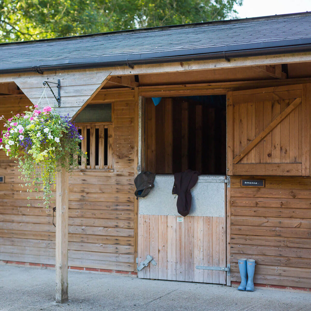 a stable block decorated by a hanging basket full of wild flowers in bloom with a coat hanging over a stable door and a pair of blue wellington boots outside on the floor