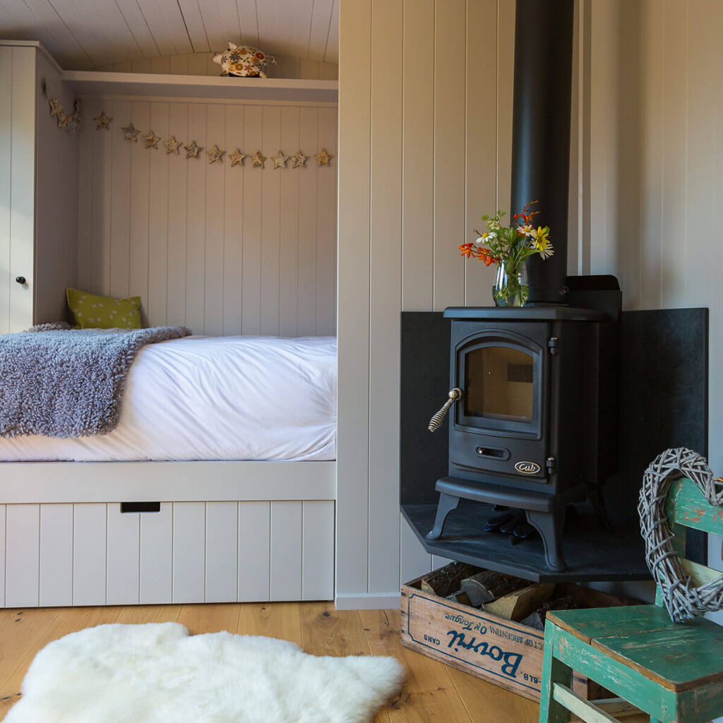 Inside one of the beautifully fitted out shepherd huts at Settledown retreats. A view of the wood burning stove, sheepskin rug on the floor, king size bed in the background