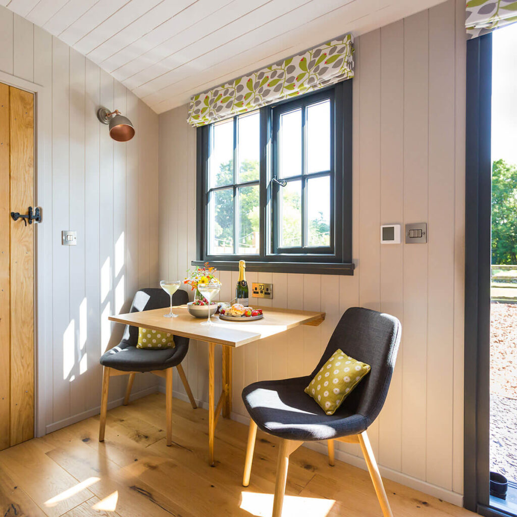 sun streams through the window of a lovely shepherd hut, a bottle of Champagne, food and wild flowers on the table