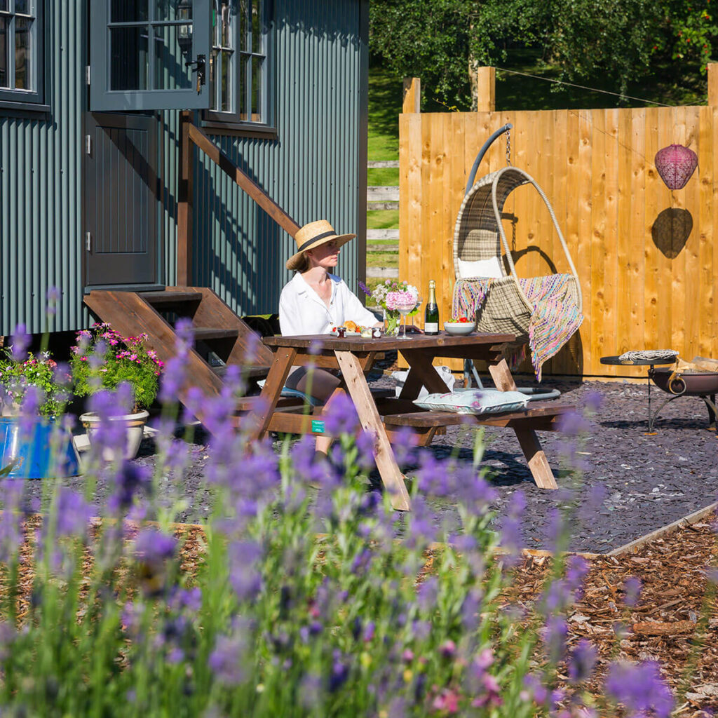 a woman with straw sun hat relaxes in the sun sat at a patio table outside a shepherd hut